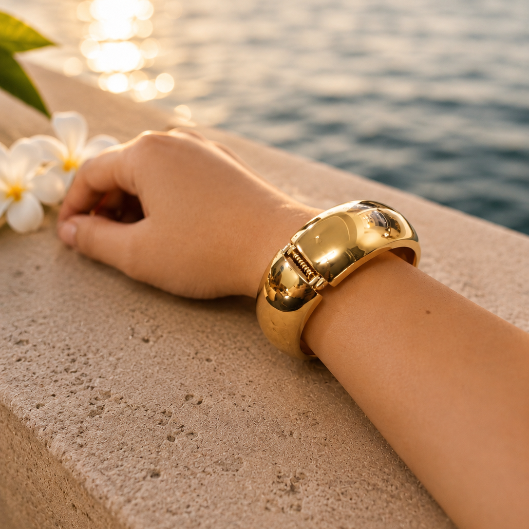 Hand wearing a gold bracelet with a sunset over water in the background