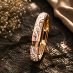 Gold ring with pink and white inlay on a dark surface with flowers in the background