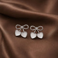 Pair of silver earrings with heart and bow design on a brown fabric background