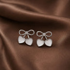 Pair of silver earrings with heart and bow design on a brown fabric background