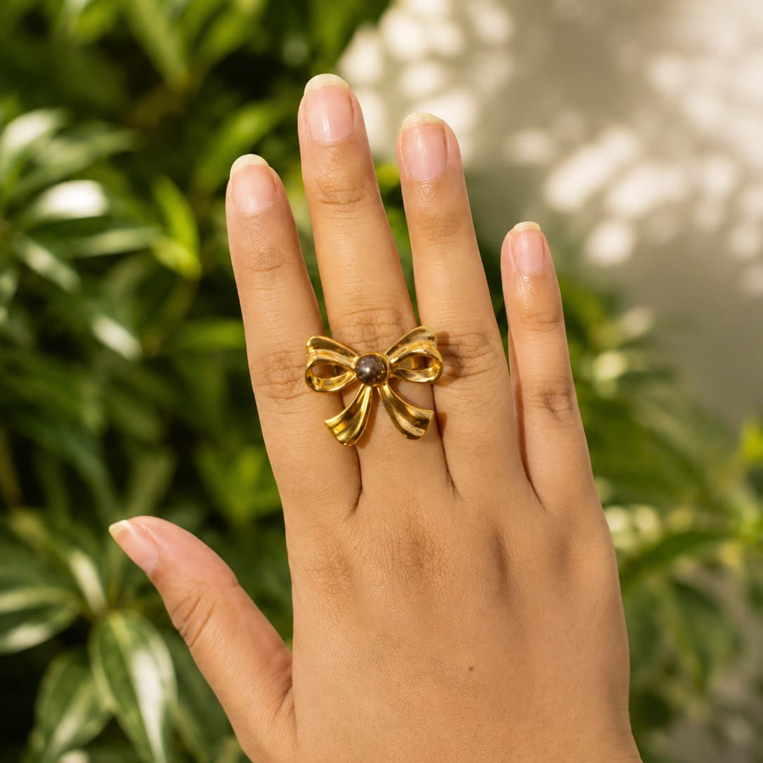 Hand wearing a gold bow-shaped ring with a green leafy background