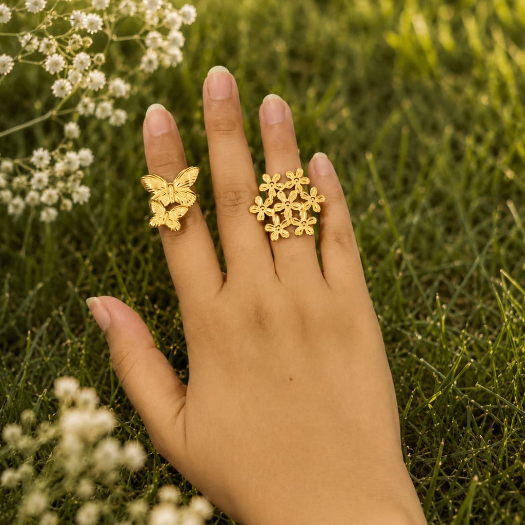 Hand wearing two gold rings with floral designs against a natural background of grass and small white flowers.