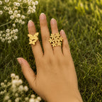 Hand wearing two gold rings with floral designs against a natural background of grass and small white flowers.