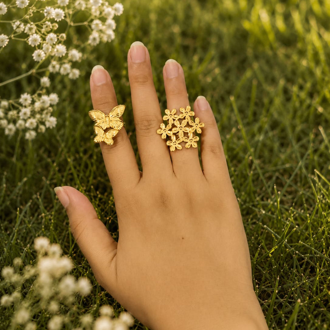 Hand wearing two gold rings with floral designs against a natural background of grass and small white flowers.