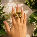 Hand wearing a gold ring with floral design, surrounded by greenery and flowers.