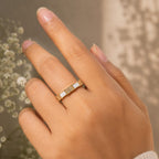 Hand wearing a gold ring with a white stone against a blurred natural background