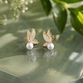 Pair of earrings with gold butterfly wings and pearl centers on a green surface with leaves in the background.