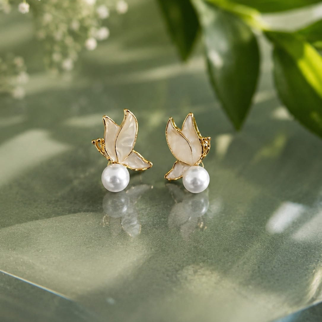 Pair of earrings with gold butterfly wings and pearl centers on a green surface with leaves in the background.