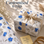 Gift wrapped in white paper with blue floral designs, gold ribbon, and a gold card on a light surface.