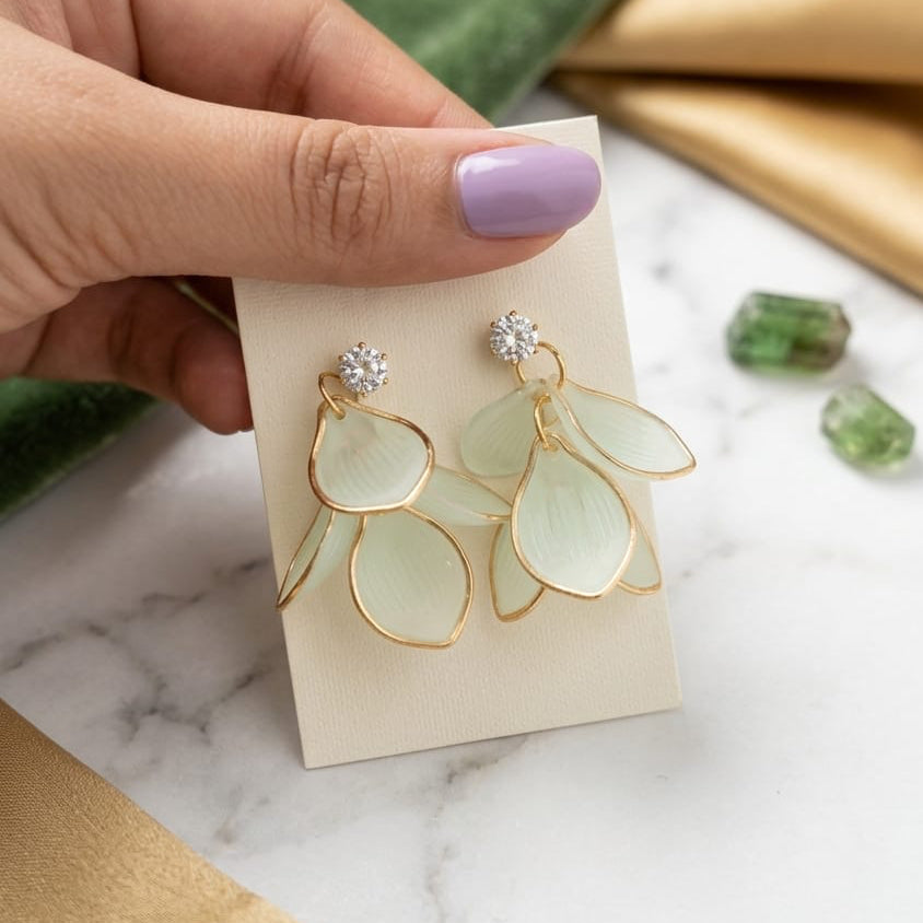 Hand holding a pair of earrings on a marble surface with green and gold elements.
