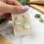 Hand holding a pair of earrings on a marble surface with green and gold elements.